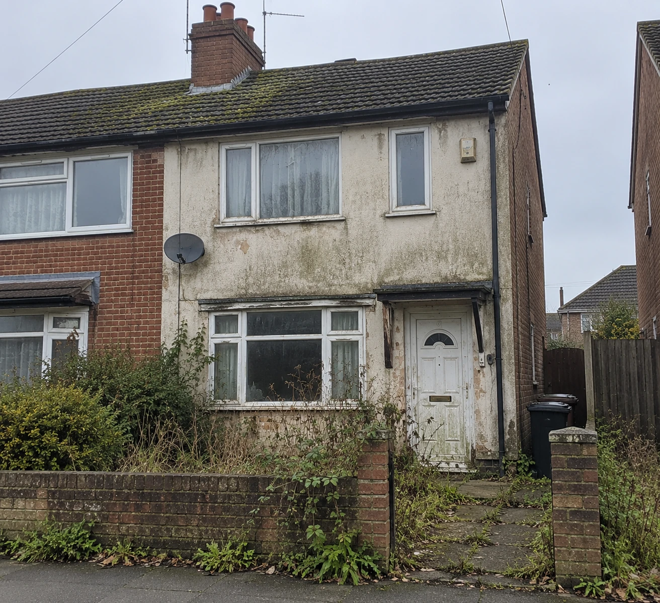 A neglected semi-detached house seen from the pavement, its render stained and peeling, moss across the roof tiles, brambles growing through the front garden wall and the front door weathered bare