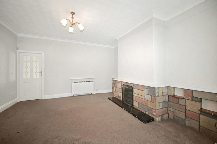 The front reception room, empty, with a stone clad chimney breast, a back boiler behind the hearth and worn brown carpet throughout