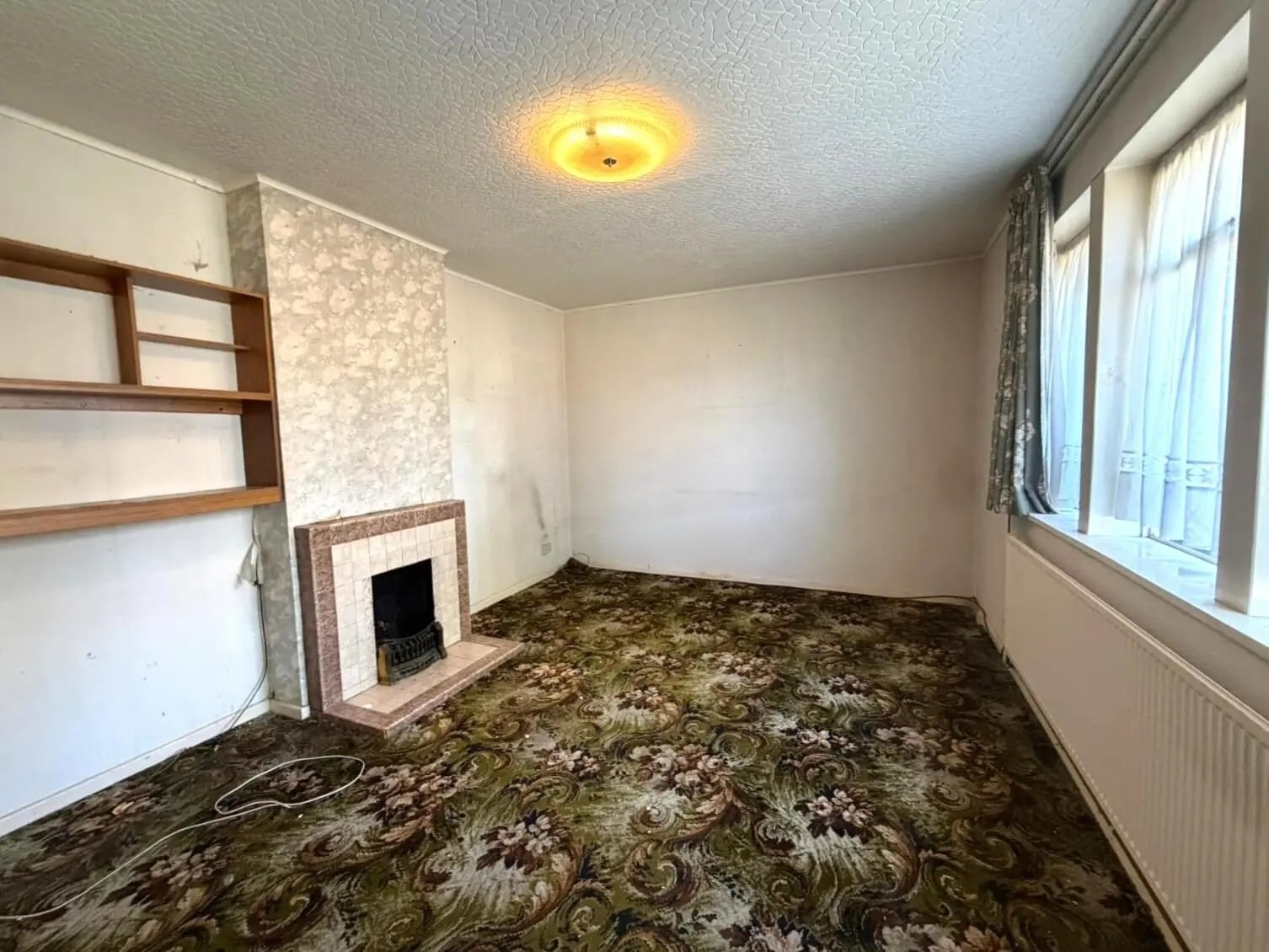 The rear reception room, with a tiled fireplace, built in shelving and a patterned green carpet, damp marking the wall beside the chimney