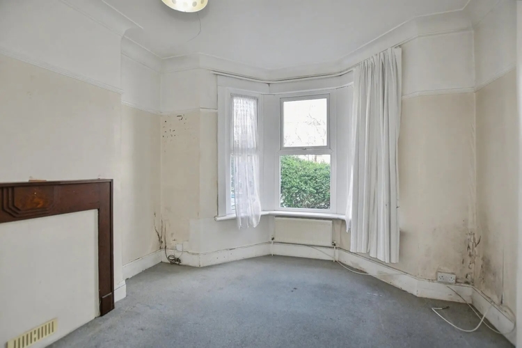 The front room, with damp staining running along the skirting beneath the bay window and cabling left loose across the carpet